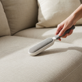 Gros plan montrant une brosse anti-poils rose en action, séparant un tas de poils de chat agglomérés d'une surface de canapé en velours rose, illustrant l'efficacité du nettoyage.